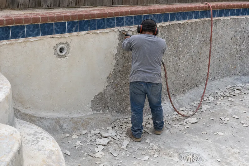 Worker with a chipping hammer stripping old plaster from a pool wall