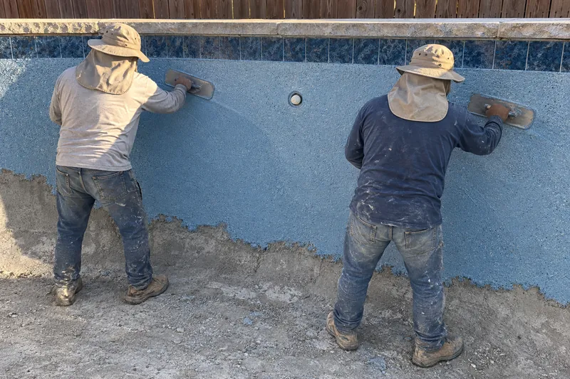 Two finishers hand-troweling speckled quartz plaster in an empty pool